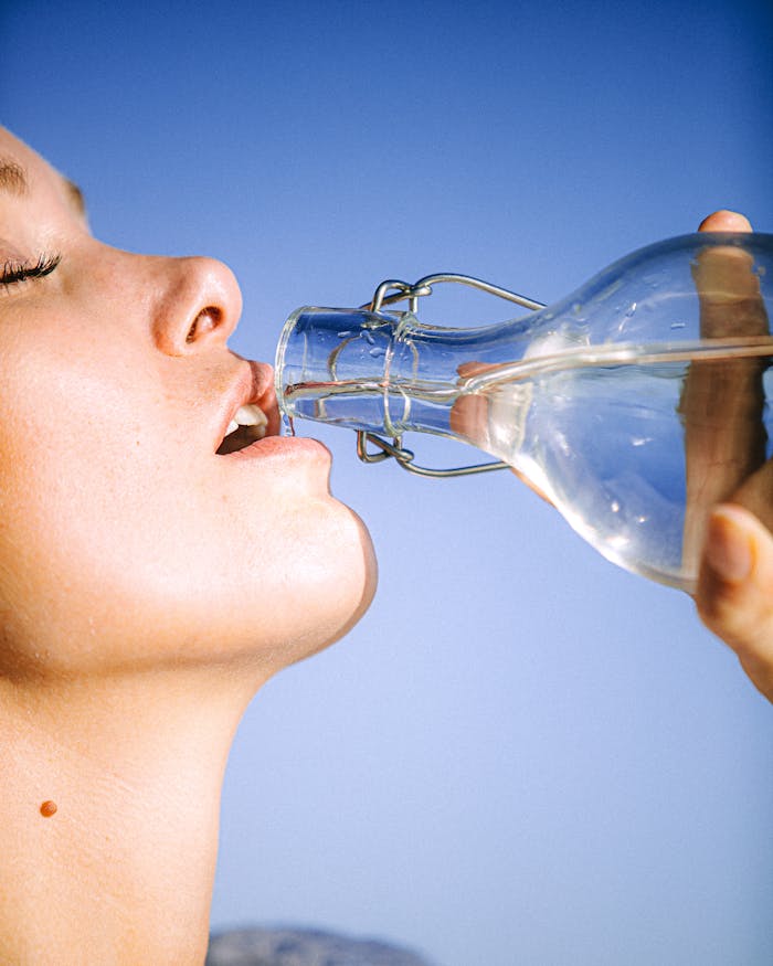 The Art of Drawing Readers In: Your attractive post title goes here A woman drinking water from a glass bottle against a clear blue sky, emphasizing hydration.